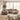 A plush, modular upholstered sectional sofa in a warm taupe-beige color, styled in a bright, modern living room with sheer curtains, a large woven area rug, and a potted olive tree.