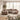 A plush, modular upholstered sectional sofa in a warm taupe-beige color, styled in a bright, modern living room with sheer curtains, a large woven area rug, and a potted olive tree.
