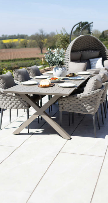 Outdoor dining set on a light stone patio featuring a modern beige garden table and chairs set, alongside a large double egg chair. The scene is set in a garden with fields and trees in the background under a clear blue sky.