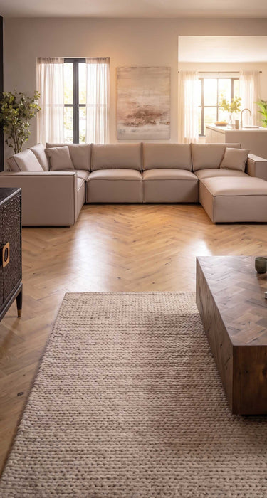 A large modular leather corner sofa with a chaise lounge in a modern, sunlit living room with herringbone wood floors and a textured jute rug.