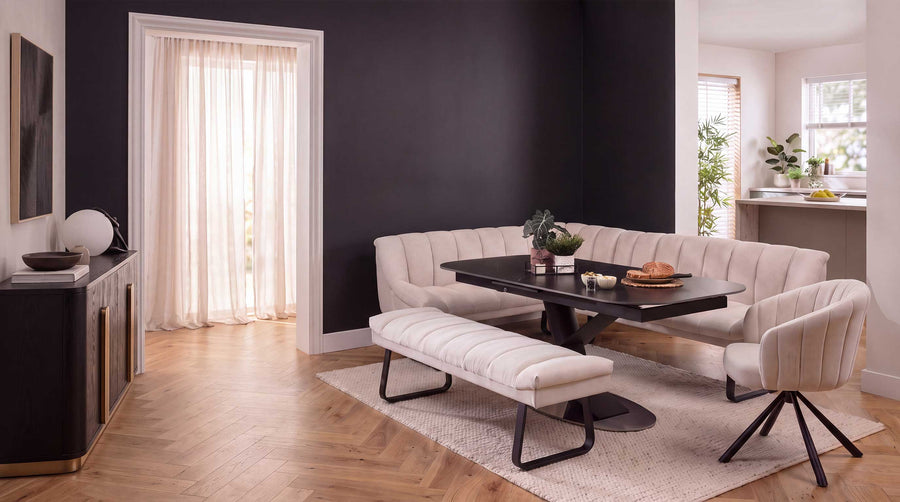 A modern dining room setup featuring a cream tufted corner dining bench, a matching backless bench, a black ceramic marble dining table, and a cream swivel chair on a light rug, set against a dark charcoal wall and light wood flooring.