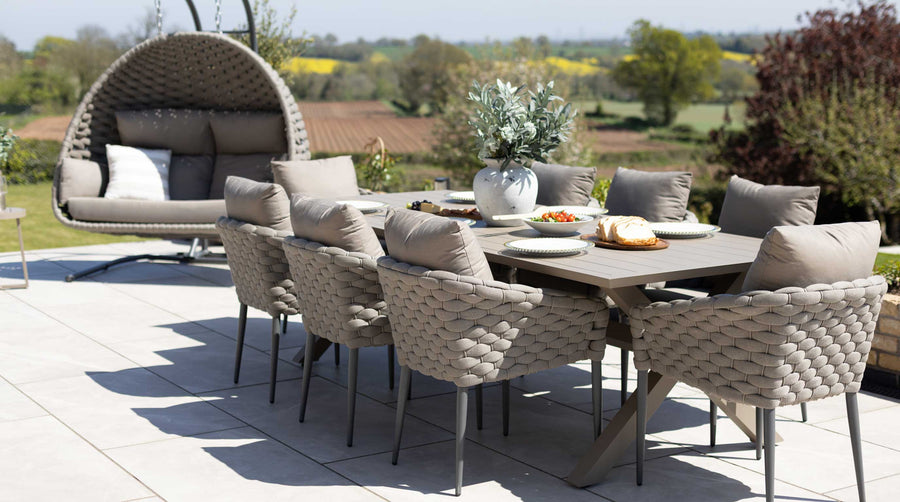 Outdoor dining set on a patio featuring a large 8 seater garden table and chair set, and a hanging egg chair in the background, set against a scenic rural landscape.